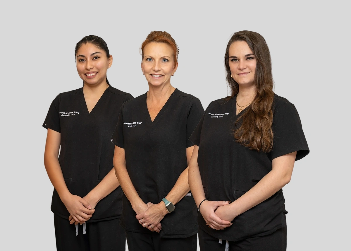Three female dental professionals in black scrubs standing together against gray background.