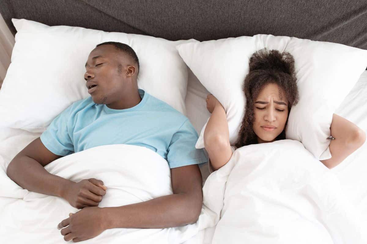 Man snoring in bed while woman lies awake, illustrating sleep apnea symptoms requiring dental treatment.